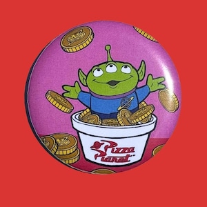 May include: A round button featuring a green alien character with three eyes, wearing a blue shirt, and surrounded by gold coins. The alien is in a white bowl filled with coins, with the words "Pizza Planet" on the bowl.