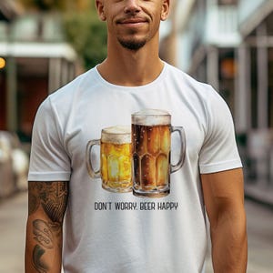 Puede incluir: Camiseta blanca con un gráfico de dos jarras de cerveza. El texto "Don't Worry. Beer Happy" está impreso debajo de las jarras.