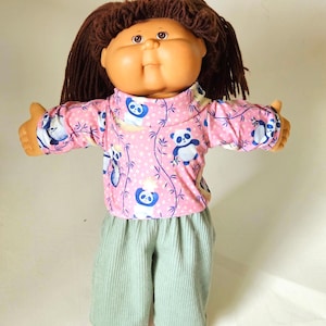 May include: A doll with a round face, tan skin, and brown yarn hair. The doll wears a pink long-sleeved shirt featuring a repeating pattern of blue and white pandas and bamboo stalks, paired with light green corduroy pants and light pink shoes.