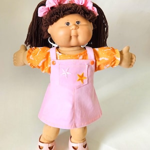 Könnte beinhalten: Eine Puppe mit braunem Wollhaar, das zu zwei Pferdeschwänzen mit rosa Haargummis gebunden ist. Die Puppe trägt ein orangefarbenes kurzärmeliges Oberteil mit weißem Muster, ein hellrosa Latzkleid mit zwei gestickten Sternen und rosa Schuhe.