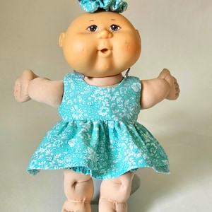 Peut inclure: Une poupée au visage rond, aux yeux marron foncé et à la petite touffe de cheveux noirs attachée avec un chouchou à motif floral bleu sarcelle. La poupée porte une robe sans manches bleu sarcelle à motif floral blanc et une culotte assortie.