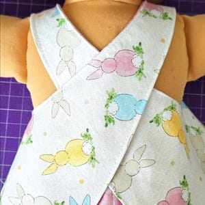 May include: A doll wearing a white apron with a colourful bunny print. The bunnies are in pastel colours of pink, blue, and yellow, with green leafy accents. The doll's outfit includes a pink polka-dot skirt.