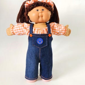 May include: A vintage doll with brown hair and a peach-colored face. The doll wears a white and orange gingham shirt, denim dungarees with a smiley face patch, and a matching hair bow. The doll has brown eyes and is wearing hoop earrings.
