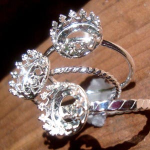May include: Close-up of several silver-colored crown rings. The rings have intricate filigree detailing on the crown and band. The rings are displayed on a wooden surface, with shadows adding depth.