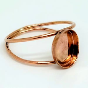 May include: A copper-colored ring with an oval setting. The ring's band has a double-loop design, with the setting positioned on top. The ring is made of a shiny, metallic material.