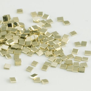 Könnte beinhalten: Ein Haufen kleiner, quadratischer Metallstücke in Goldfarbe. Die Stücke sind auf einer weißen Oberfläche verstreut.