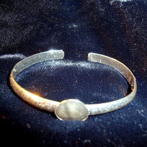 Op de afbeelding: Een zilveren manchetarmband met een ovaalvormige middensteen. De armband heeft een open ontwerp en een gehamerde textuur. Het metaal heeft een licht aangeslagen uiterlijk, waardoor het een vintage uitstraling heeft. De armband wordt gepresenteerd op een donkerblauwe fluwelen ondergrond.