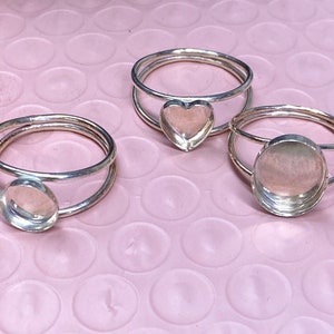 May include: Three silver rings with different shaped settings. The rings are double-banded and have a round, heart, and oval shaped setting.