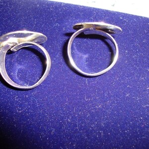 May include: Two silver rings with oval-shaped tops. The rings are on a blue velvet surface.