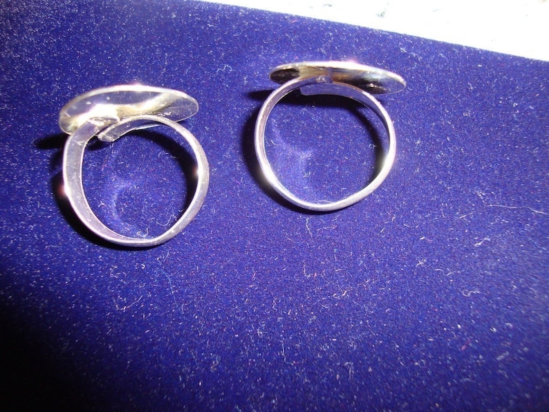 May include: Two silver rings with oval-shaped tops. The rings are on a blue velvet surface.