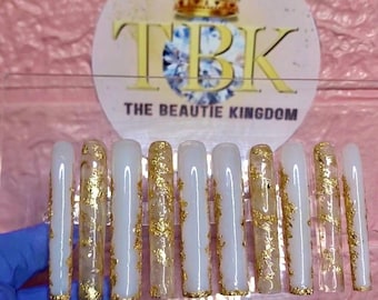 May include: A set of long, square-shaped artificial nails. Each nail is painted white with gold foil accents. The nails are displayed in front of a pink background with a logo that reads "TBK THE BEAUTIE KINGDOM".