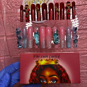 May include: A display of artificial nails in various designs. Some nails are a deep red with gold accents, others are pink with glitter and teal designs, and some are blue with crystal embellishments. A box with the text "The Lash Castle" is also visible.