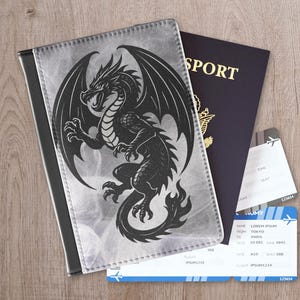 May include: A black and gray dragon design passport holder with a black border. A dark blue passport and two airline tickets are next to the passport holder on a wooden surface. The tickets have the text "LOREM IPSUM" and "TOKYO TO PARIS".