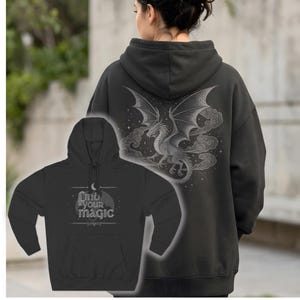 May include: Dark gray hoodie featuring a detailed dragon graphic on the back with wings spread, clouds, and stars. The front shows a smaller hoodie with the text "Find your magic" and a crescent moon design. The hoodie has a drawstring hood and a front pocket.