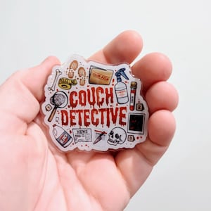 May include: A clear, irregular-shaped sticker with the words "COUCH DETECTIVE" in red, surrounded by detective-themed illustrations. The illustrations include a magnifying glass, footprints, a skull, and a file folder.