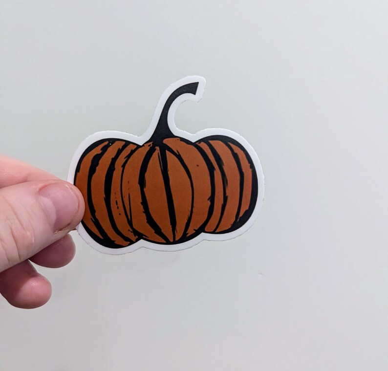 May include: A sticker featuring a stylized orange pumpkin with black vertical lines and a curved stem. The pumpkin has a white border. The sticker is held by a hand against a white background. Perfect for fall and Halloween.