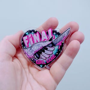 May include: A heart-shaped sticker with the words "FINAL GIRL GANG" and a hand holding a knife. The design features a black and pink color scheme with a turquoise background and stars.
