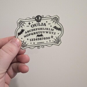 K&ouml;nnte beinhalten: Ein cremefarbener Ouija-Brett-Aufkleber mit schwarzer Schrift und Grafiken. Das Design umfasst das Alphabet, Zahlen und die W&ouml;rter "YES", "NO" und "GOODBYE". Dekorative Elemente sind Flederm&auml;use, Spinnweben und ein Totenkopf.