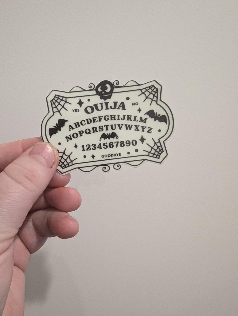 K&ouml;nnte beinhalten: Ein cremefarbener Ouija-Brett-Aufkleber mit schwarzer Schrift und Grafiken. Das Design umfasst das Alphabet, Zahlen und die W&ouml;rter "YES", "NO" und "GOODBYE". Dekorative Elemente sind Flederm&auml;use, Spinnweben und ein Totenkopf.
