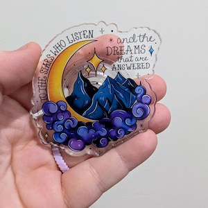 May include: A clear acrylic sticker featuring a crescent moon, mountains, stars, and clouds in shades of yellow, blue, and purple. The sticker has the text "TO THE STARS WHO LISTEN" and "and the DREAMS that are ANSWERED".