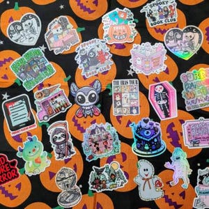 May include: A collection of Halloween-themed stickers on a black and orange pumpkin-patterned background. The stickers feature various spooky designs, including ghosts, bats, and skeletons, with phrases like "Read More Horror" and "Spooky Book Club".