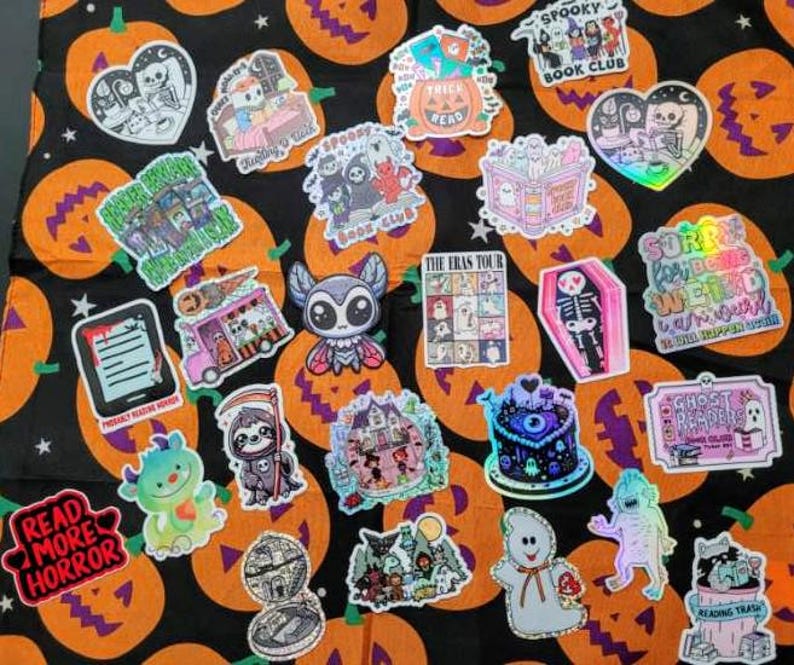 May include: A collection of Halloween-themed stickers on a black and orange pumpkin-patterned background. The stickers feature various spooky designs, including ghosts, bats, and skeletons, with phrases like "Read More Horror" and "Spooky Book Club".