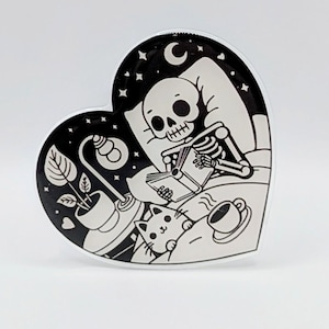 May include: Heart-shaped black and white sticker featuring a skeleton reading in bed with a cat, lamp, and coffee cup. The background is a starry night scene with a crescent moon. The sticker has a glossy finish.