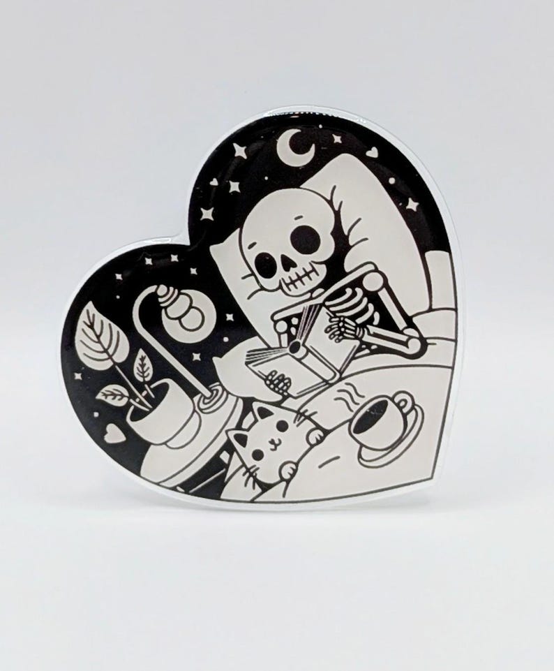 May include: Heart-shaped black and white sticker featuring a skeleton reading in bed with a cat, lamp, and coffee cup. The background is a starry night scene with a crescent moon. The sticker has a glossy finish.