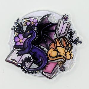 May include: A fantasy-themed sticker featuring two dragons, one purple and one gold, with open books and purple flowers. The purple dragon has black wings. The sticker has a clear, iridescent border and is set against a white background.