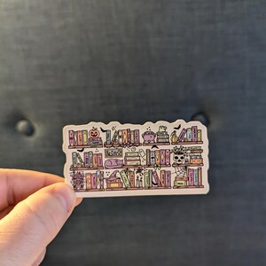 May include: A sticker featuring a whimsical bookshelf design with Halloween-themed elements. The shelves are filled with colorful books, a pumpkin, a cauldron, a ghost, and a skull. The sticker has a white border and is held by a hand.