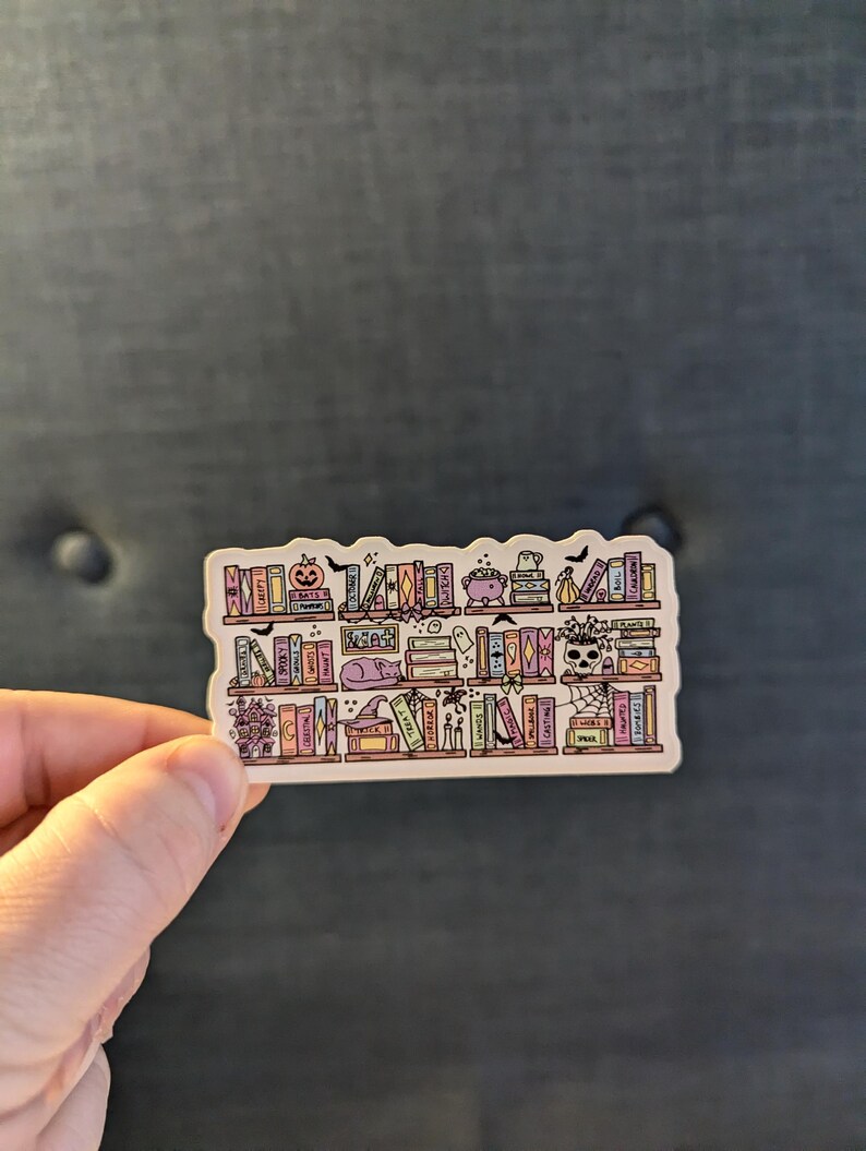 May include: A sticker featuring a whimsical bookshelf design with Halloween-themed elements. The shelves are filled with colorful books, a pumpkin, a cauldron, a ghost, and a skull. The sticker has a white border and is held by a hand.