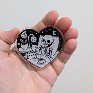 May include: A heart-shaped acrylic charm featuring a black and white illustration of a skeleton reading in bed with a cat and a cup of coffee. The design includes a night scene with stars, a moon, a lamp, and a potted plant.