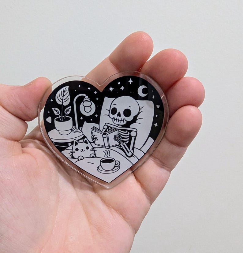 May include: A heart-shaped acrylic charm featuring a black and white illustration of a skeleton reading in bed with a cat and a cup of coffee. The design includes a night scene with stars, a moon, a lamp, and a potted plant.