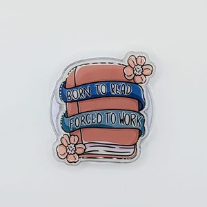 May include: A clear, rounded sticker featuring a book with the text "BORN TO READ FORCED TO WORK" on blue banners. Pink flowers are on either side of the book. The book is a reddish-brown color.