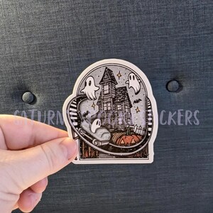 K&ouml;nnte beinhalten: Ein Aufkleber mit einem Spukhaus mit Geistern, Flederm&auml;usen und einer Achterbahnstrecke. Vor dem Haus steht ein K&uuml;rbis. Der Aufkleber hat den Text "SATURN STICKERS" auf der Oberseite.