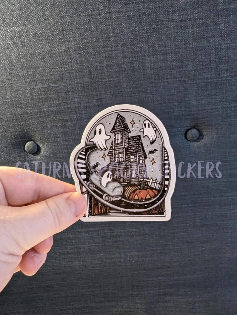 K&ouml;nnte beinhalten: Ein Aufkleber mit einem Spukhaus mit Geistern, Flederm&auml;usen und einer Achterbahnstrecke. Vor dem Haus steht ein K&uuml;rbis. Der Aufkleber hat den Text "SATURN STICKERS" auf der Oberseite.