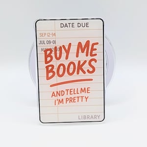 May include: A rectangular library card-themed sticker with the text "BUY ME BOOKS AND TELL ME I'M PRETTY" in red. The sticker has a white background with a lined paper design and the word "LIBRARY" at the bottom.