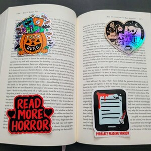 May include: Open book with four horror-themed stickers. One sticker says "Trick or Read" on a pumpkin. Another says "Read More Horror". The third shows a skeleton reading. The last shows a tablet with blood and the text "Probably Reading Horror".