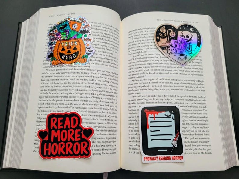 May include: Open book with four horror-themed stickers. One sticker says "Trick or Read" on a pumpkin. Another says "Read More Horror". The third shows a skeleton reading. The last shows a tablet with blood and the text "Probably Reading Horror".