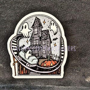 K&ouml;nnte beinhalten: Ein Halloween-Sticker mit einem Spukhaus, Geistern, einem K&uuml;rbis und einem Achterbahnwagen mit einem Geisterpassagier. Der Aufkleber hat ein Schwarz-Wei&szlig;-Farbschema mit orangefarbenen und grauen Akzenten, und der Text "Happy Halloween" ist sichtbar.