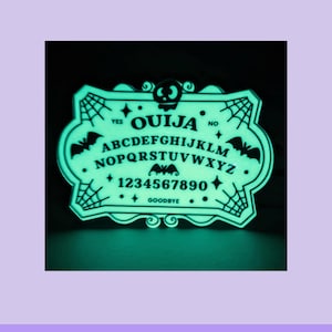 K&ouml;nnte beinhalten: Ein leuchtend gr&uuml;ner Ouija-Brett-Aufkleber mit schwarzem Rand. Das Brett zeigt das Alphabet, Zahlen und die W&ouml;rter "Ja", "Nein" und "Auf Wiedersehen". Flederm&auml;use und Spinnweben verzieren das Design. Der Text "WATERPROOF VINYL STICKER" steht unten.