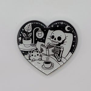 May include: Heart-shaped enamel pin with a black and white illustration. A skeleton reads in bed with a cat, a cup of coffee, and a lamp. The pin has a silver border and a night sky design.