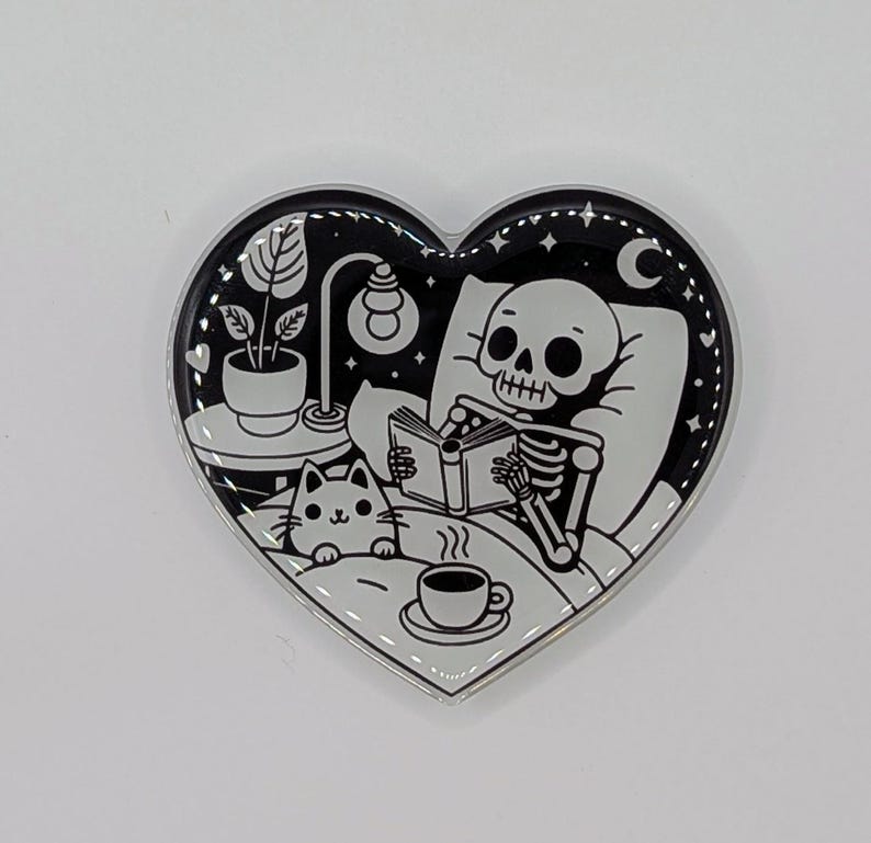 May include: Heart-shaped enamel pin with a black and white illustration. A skeleton reads in bed with a cat, a cup of coffee, and a lamp. The pin has a silver border and a night sky design.
