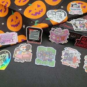 May include: A collection of Halloween-themed stickers featuring spooky illustrations. Designs include ghosts, skeletons, pumpkins, and text like "Read More Horror" and "Spooky Book Club." The stickers are on a black surface with a Halloween-themed fabric.