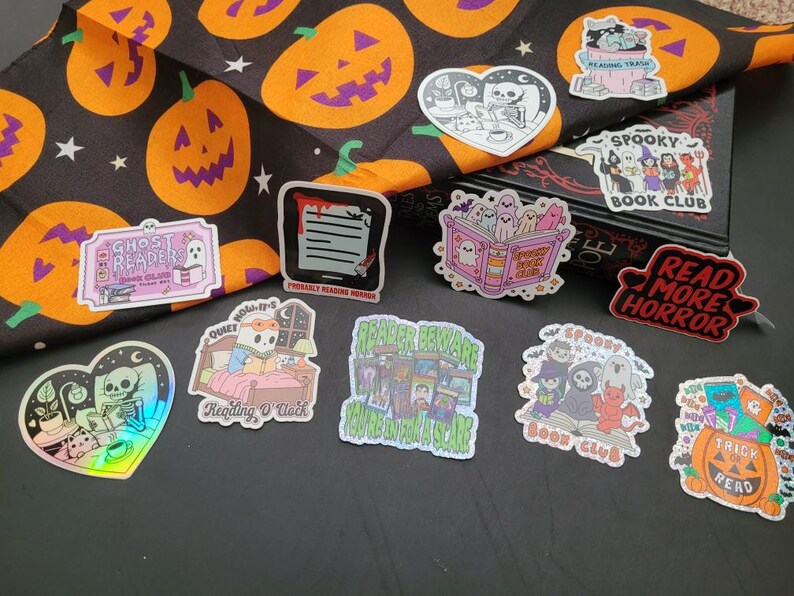 May include: A collection of Halloween-themed stickers featuring spooky illustrations. Designs include ghosts, skeletons, pumpkins, and text like "Read More Horror" and "Spooky Book Club." The stickers are on a black surface with a Halloween-themed fabric.
