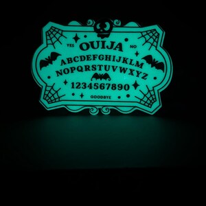 K&ouml;nnte beinhalten: Ein leuchtend gr&uuml;nes Ouija-Brett mit schwarzer Schrift und dekorativen Elementen. Das Brett zeigt das Alphabet, Zahlen und die W&ouml;rter "Ja", "Nein" und "Auf Wiedersehen". Flederm&auml;use und Spinnweben schm&uuml;cken die R&auml;nder und erzeugen eine gruselige &Auml;sthetik.