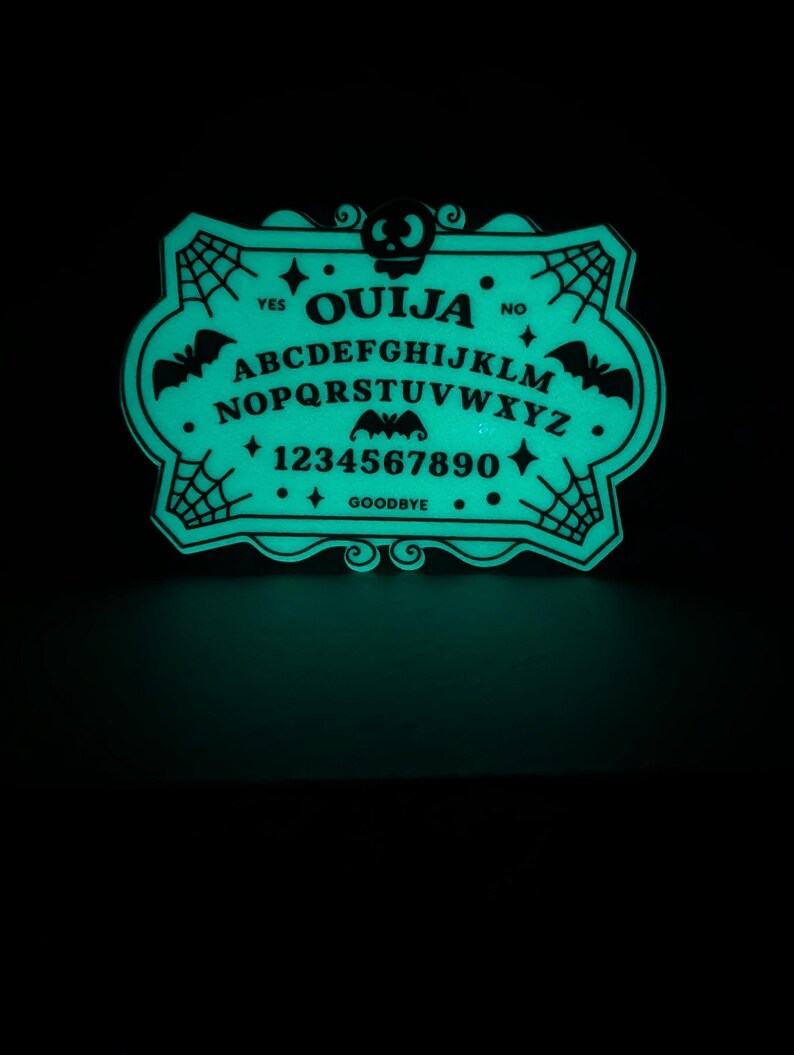 K&ouml;nnte beinhalten: Ein leuchtend gr&uuml;nes Ouija-Brett mit schwarzer Schrift und dekorativen Elementen. Das Brett zeigt das Alphabet, Zahlen und die W&ouml;rter "Ja", "Nein" und "Auf Wiedersehen". Flederm&auml;use und Spinnweben schm&uuml;cken die R&auml;nder und erzeugen eine gruselige &Auml;sthetik.