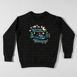 May include: Black sweatshirt with a whimsical graphic. The design features a blue car with passengers, trees, stars, and a crescent moon. Text reads "Get in loser, We're going to Therapy."