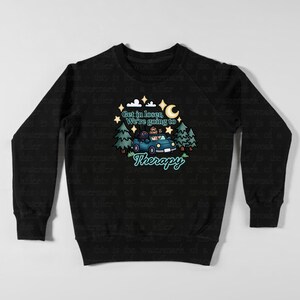 May include: Black sweatshirt with a whimsical graphic. The design features a blue car with passengers, trees, stars, and a crescent moon. Text reads "Get in loser, We're going to Therapy."