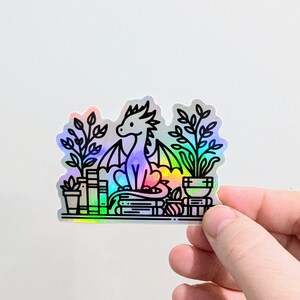 May include: A holographic sticker featuring a dragon sitting on books, surrounded by potted plants. The dragon has a rainbow-colored body and black outlines. The sticker is held by a hand against a white background.